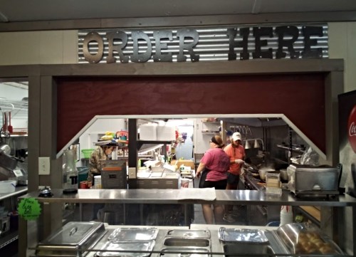 A food counter with a sign reading "ORDER HERE," showing staff preparing meals in a kitchen behind.