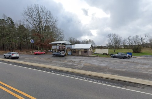 A small roadside building with a sign, surrounded by trees and parked cars, under a cloudy sky.