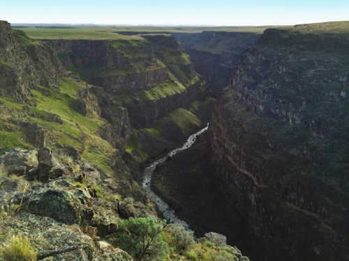 A scenic view of a deep canyon with a winding river at the bottom, surrounded by lush green vegetation and rocky cliffs.