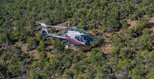 A helicopter flying over a dense forested area with green trees and rocky terrain below.