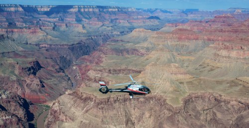 A helicopter flies over the Grand Canyon, showcasing its vast, colorful rock formations and deep valleys.