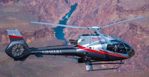 A helicopter flies over a canyon, with a river visible below and rugged terrain in the background.