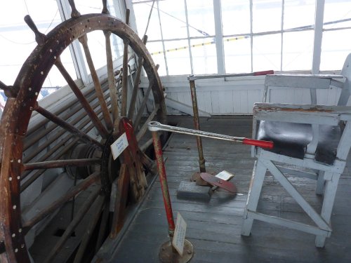 A vintage ship's wheel and control levers in a nautical setting, with wooden chairs and tools in the background.