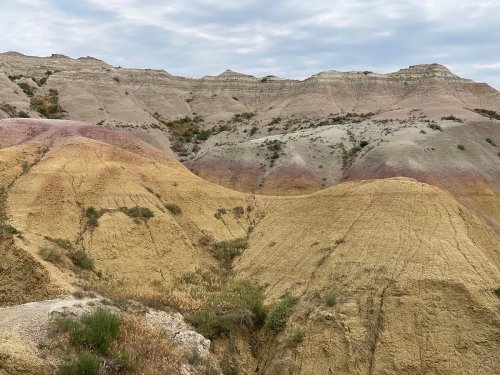 Colorful, layered hills with shades of yellow, pink, and gray under a cloudy sky in a natural landscape.