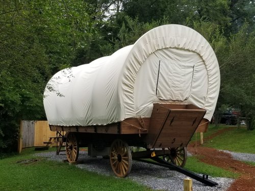 A covered wagon on wooden wheels, parked on gravel surrounded by greenery.