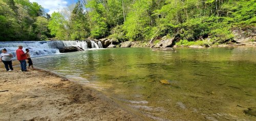A serene river scene with a waterfall, surrounded by lush greenery and people enjoying the sandy shore.