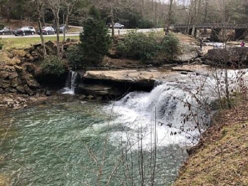 A serene waterfall flows into a clear pool, surrounded by trees and rocky terrain, with a road and cars in the background.