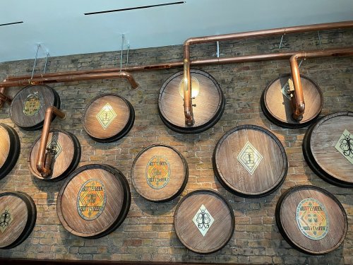A wall featuring wooden barrels with decorative labels and copper pipes against a brick background.
