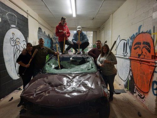 A group of five people poses around a damaged car in a graffiti-covered room, holding baseball bats and smiling.