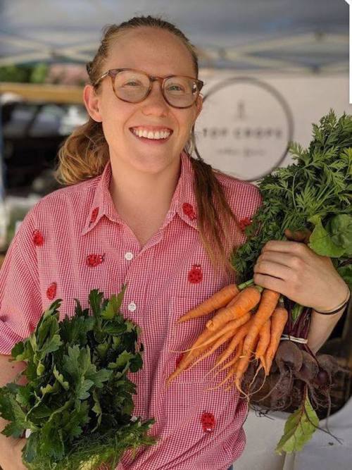 A smiling woman in a red checkered shirt holds fresh vegetables, including carrots and leafy greens, at a market.
