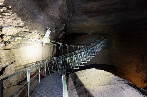 A narrow metal bridge suspended in a dark cave, illuminated by lights, leading into a rocky cavern.