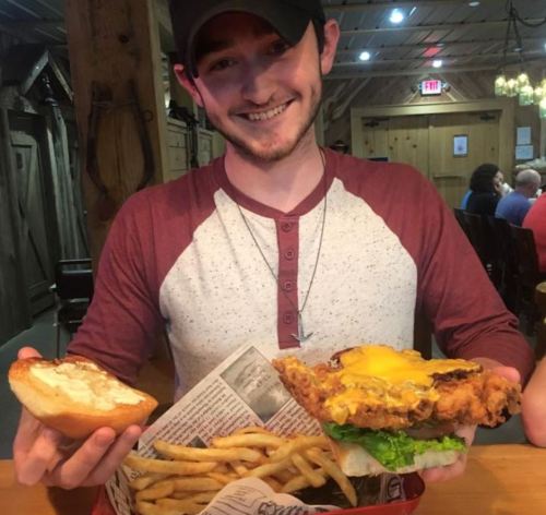 A smiling young man holds a large fried chicken sandwich with cheese and fries on a table in a casual dining setting.