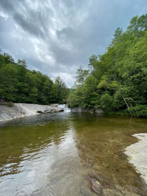 A serene river flows through a lush green forest under a cloudy sky, with smooth rocks lining the water's edge.
