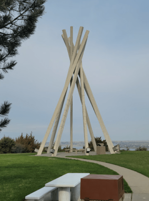 A tall, abstract sculpture made of wooden beams stands in a grassy area with a pathway and benches nearby.