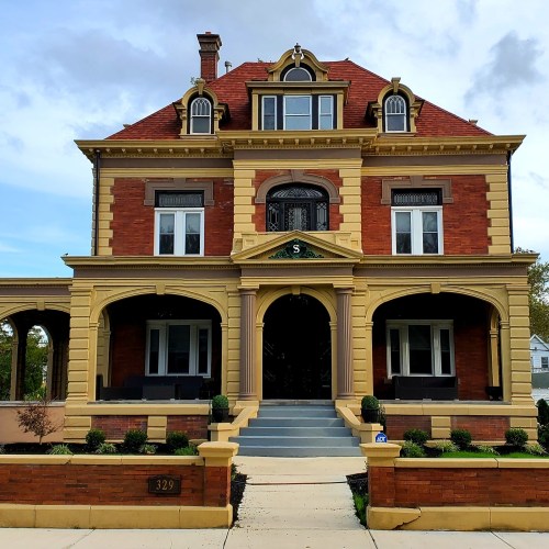 A large, ornate brick house with a red roof, arched porch, and decorative yellow trim, set against a blue sky.