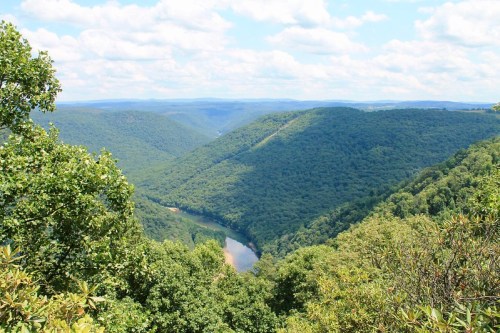 A panoramic view of lush green mountains and a winding river under a bright blue sky with fluffy clouds.