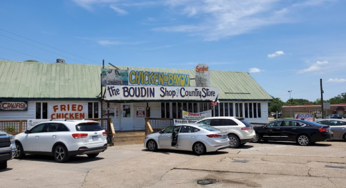 A country store with a green roof, sign reading "The Boudin Shop," and parked cars in front on a sunny day.