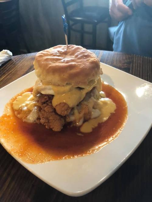 A towering biscuit sandwich with fried chicken, topped with gravy, served on a plate with a sauce pool.