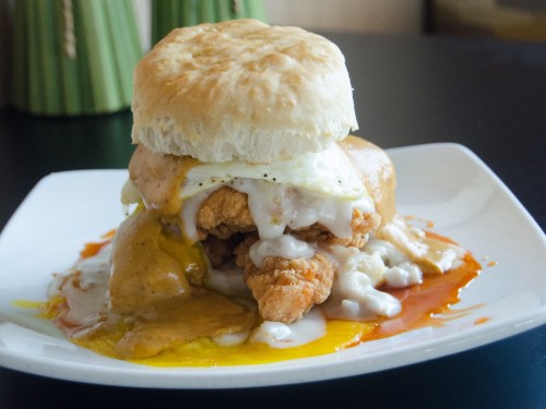 A hearty biscuit sandwich with fried chicken, a fried egg, and creamy gravy, served on a white plate.