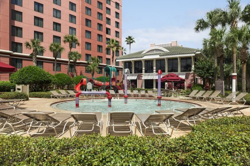 A pool area with lounge chairs, water slides, and palm trees, surrounded by greenery and a building in the background.