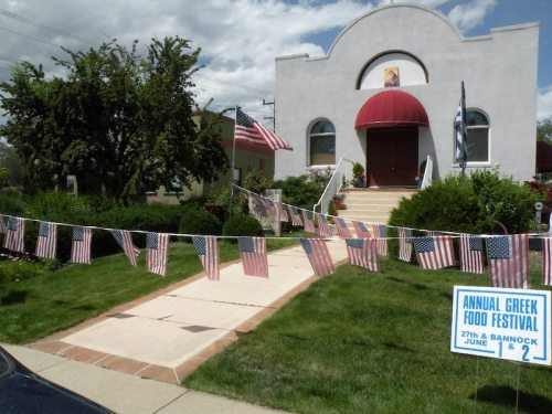 A white building with a red awning, surrounded by greenery and American flags, advertising an annual Greek food festival.