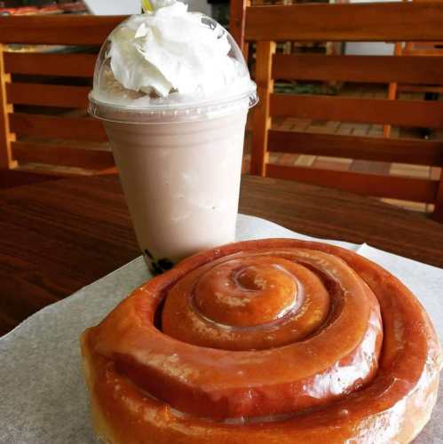 A frosty drink topped with whipped cream next to a large, glazed cinnamon roll on a table.