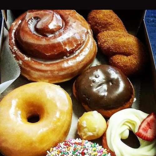 A variety of donuts including glazed, chocolate, and a cinnamon roll, arranged in a box with sprinkles and a strawberry topping.