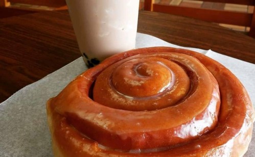 A large cinnamon roll sits on a table next to a glass of bubble tea.