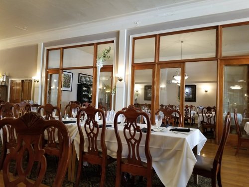 Elegant dining room with wooden chairs and tables set with white tablecloths, featuring large windows and warm lighting.