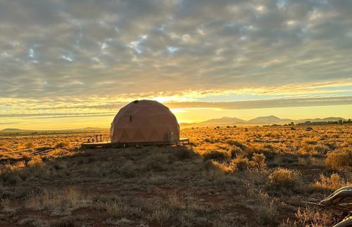 A geodesic dome home at sunrise, surrounded by a vast landscape and dramatic clouds in the sky.