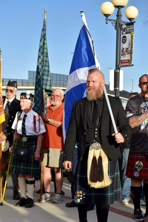 A group of people in kilts, one man holding a Scottish flag, gathered outdoors for a celebration or event.