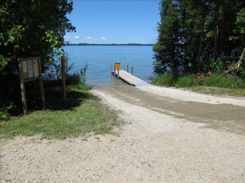 A gravel path leads to a boat ramp by a calm lake, surrounded by trees and a clear blue sky.