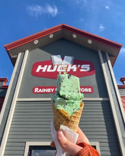 A hand holds a cone with green mint chocolate chip ice cream in front of Huck's store sign on a sunny day.