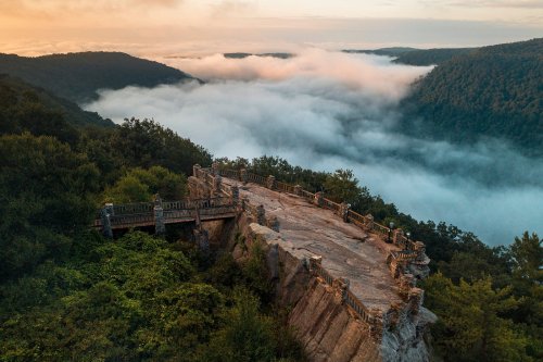 A scenic overlook with a stone platform surrounded by lush greenery and fog-covered valleys at sunrise.