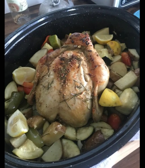 A roasted chicken surrounded by colorful vegetables and lemon slices in a black roasting pan.