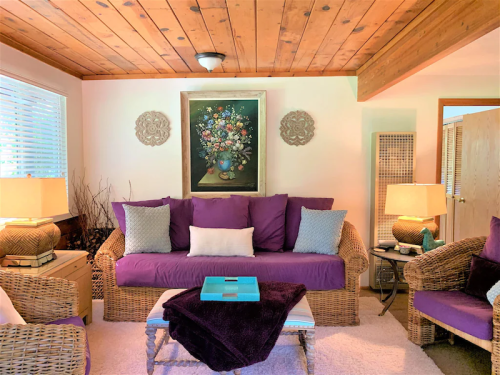 Cozy living room with purple accents, wicker furniture, a floral painting, and warm lighting.