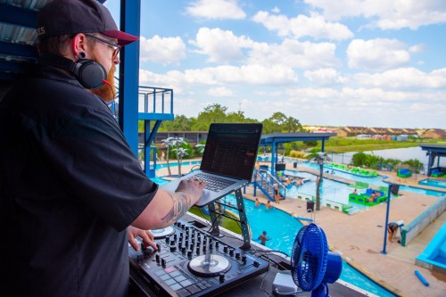 A DJ mixes music at a poolside venue, with a vibrant blue pool and people enjoying the sun in the background.