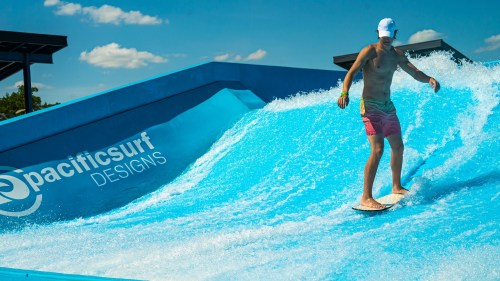A shirtless man surfing on a blue wave at a surf park, with "pacificsurf DESIGNS" logo visible on the side.