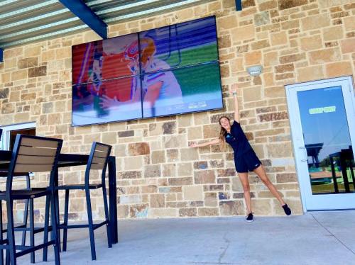 A woman stands next to a large outdoor TV displaying a sports game, with a stone wall and patio furniture in the background.