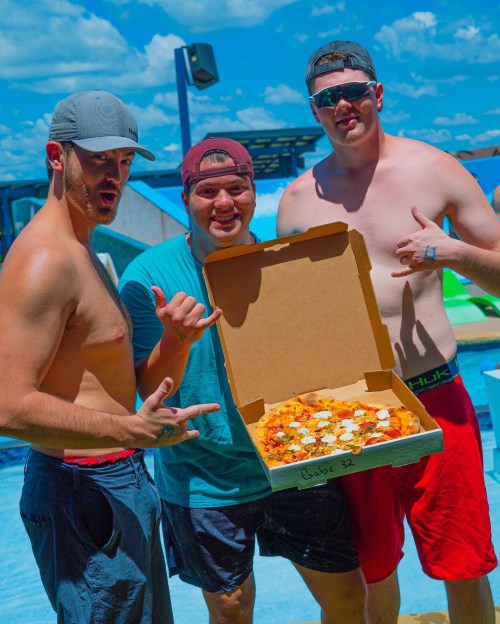 Three friends at a poolside, smiling and posing with a large pizza in a box, enjoying a sunny day.
