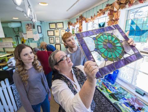 A woman shows a stained glass piece to two young adults in a bright, art-filled studio.