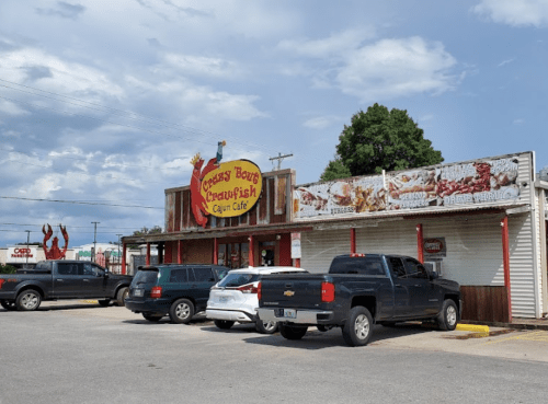 Exterior of a Cajun café named "Creepy Bows Crawfish," featuring a sign and parked vehicles in front.