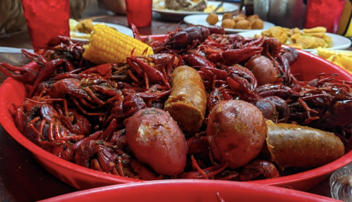 A red platter filled with crawfish, corn, sausage, and potatoes, with a blurred background of a dining setting.