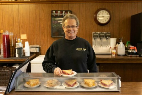 A smiling woman stands behind a counter with six slices of pie on display, in a cozy café setting.