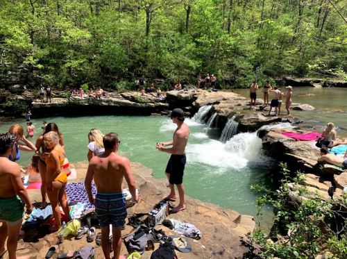 A lively swimming area with people sunbathing, swimming, and enjoying a waterfall in a lush, green setting.