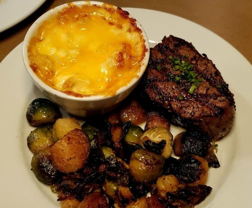A plate featuring a grilled steak, cheesy baked potatoes, and roasted Brussels sprouts with mixed vegetables.