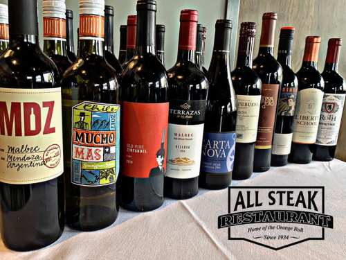 A row of wine bottles with various labels displayed on a table, featuring the All Steak Restaurant logo.