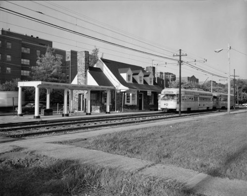Black and white image of a train station with a trolley car and surrounding buildings and greenery.