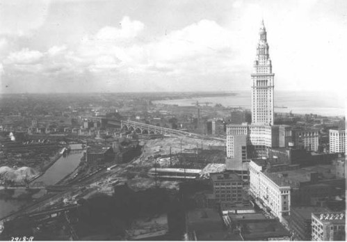 Aerial view of a cityscape featuring a tall tower, bridges, and a river, with a cloudy sky above.