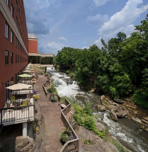 A riverside view featuring a hotel, a stone pathway, and lush greenery under a partly cloudy sky.